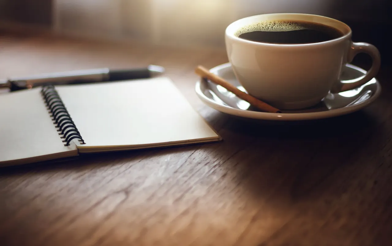 An open notebook, pen, and cup of coffee on a wooden table in warm light.