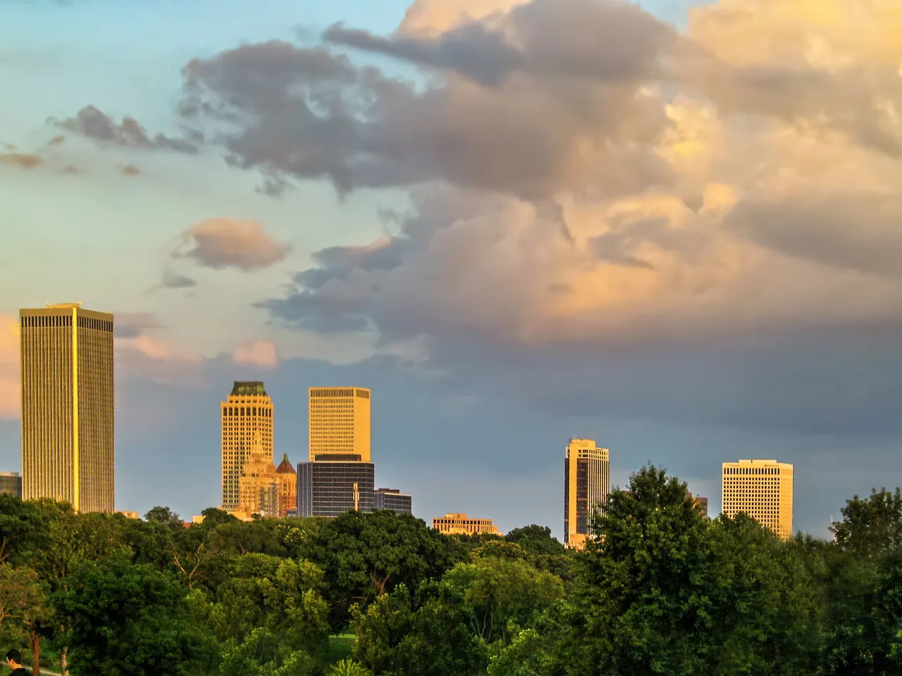 Downtown Tulsa skyline rising above green trees under a warm evening sky.
