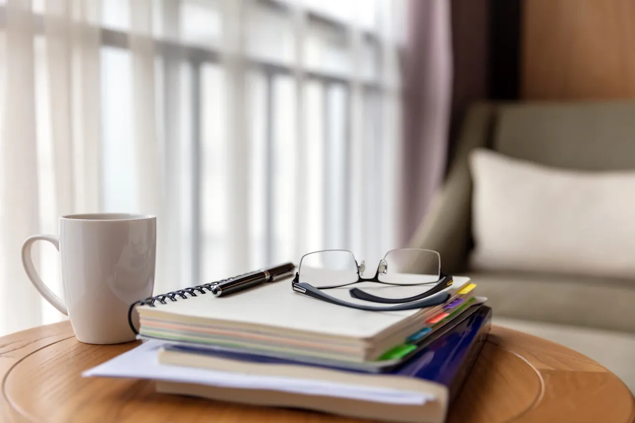 Reading glasses resting on a stack of notebooks beside a mug in a sunlit living room.